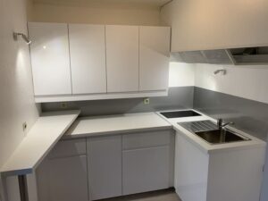 Cuisine blanche aménagée avec meubles hauts et bas, plan de travail clair, évier inox, plaques de cuisson et crédence grise.